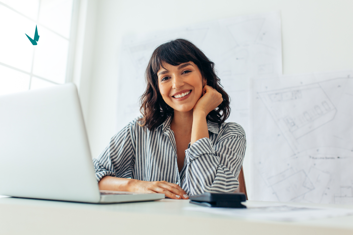 smiling woman dressed in business casual attire, sitting in front of an open latop by a window, representing the ease of working remotely from anywhere